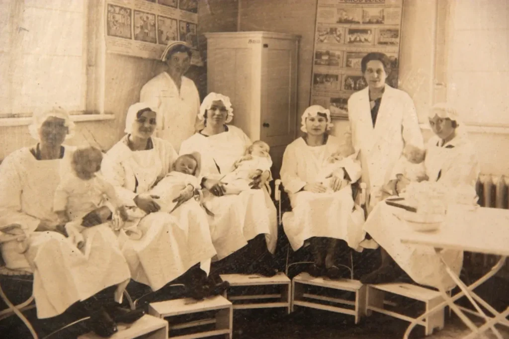 A sepia photograph of a row of women sitting in chairs and holding babies in a maternity ward, with an unknown pediatrician standing behind them on the left and Olga Rubin standing on the right